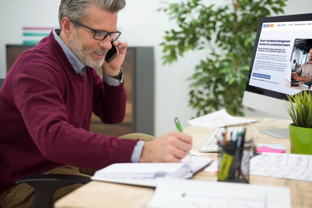 Business owner on a phone call while viewing the Bochi Web managed services page on a desktop computer, illustrating the ease of professional website support.
