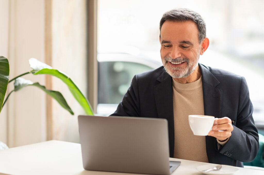 Smiling mature business owner working at a laptop with a coffee cup in a calm, bright office setting